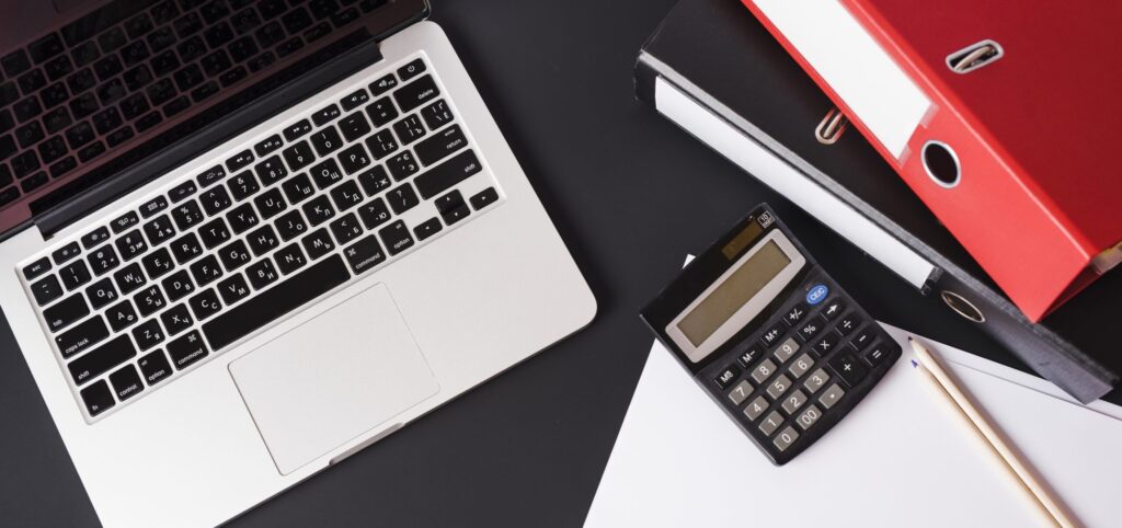 Xelous Finance & Tech. Modern workspace featuring an open laptop, a black calculator, a wooden pencil, and red and white file binders on a sleek, black office desk, symbolizing business administration and financial planning.