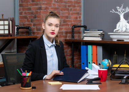 A confident young accountant Bloemfontein sits at her office desk, holding a folder. She is dressed in a professional black blazer and light blue shirt. The desk is organized with office supplies, a red coffee mug, and scattered papers, against a background of shelves with colorful binders and books. The setting reflects a modern and organized office environment.