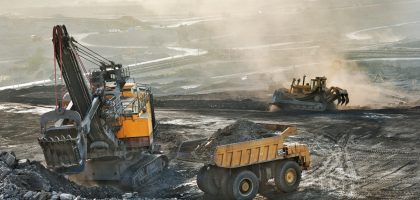 A mining site with heavy machinery in operation. A large excavator loads dirt and rocks into a dump truck, while a bulldozer works in the background amidst a dusty landscape.