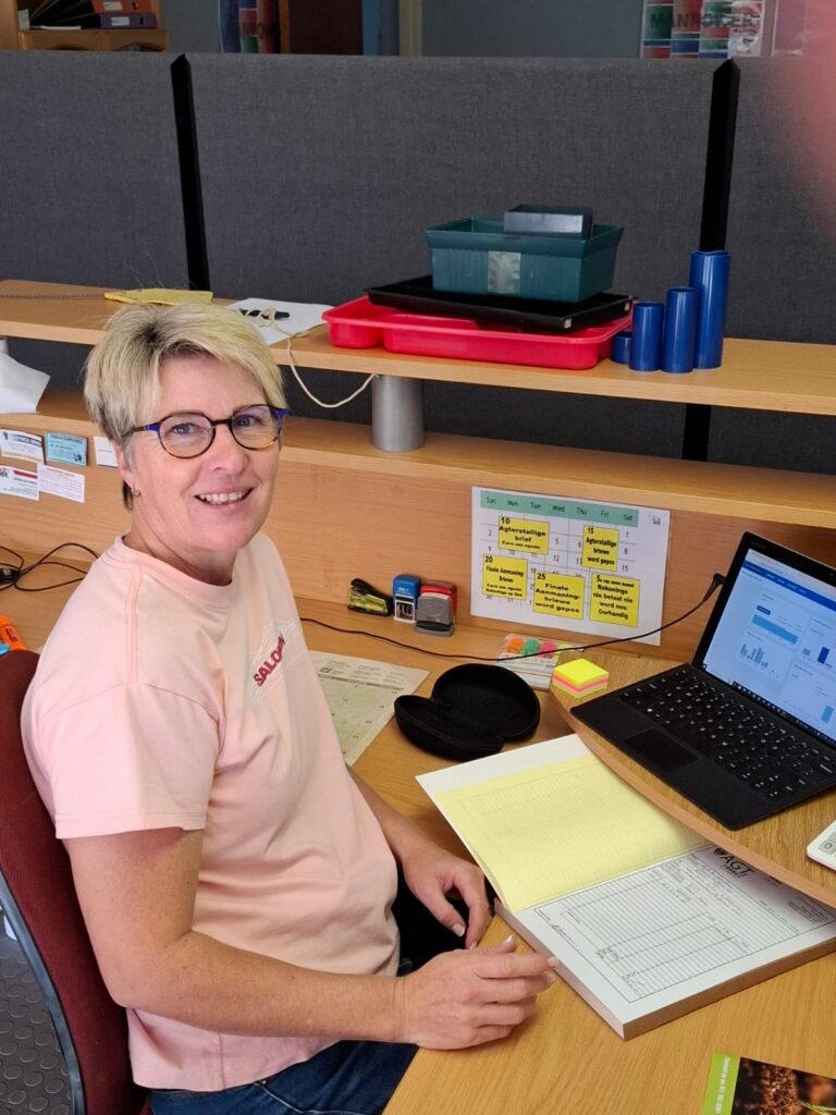 Martine Opperman is seated at her desk, busy processing information on Xero. She is wearing a light pink t-shirt and glasses, and is smiling at the camera. In front of her is an open laptop displaying financial analytics on Xero's interface, alongside various office supplies and documents.
